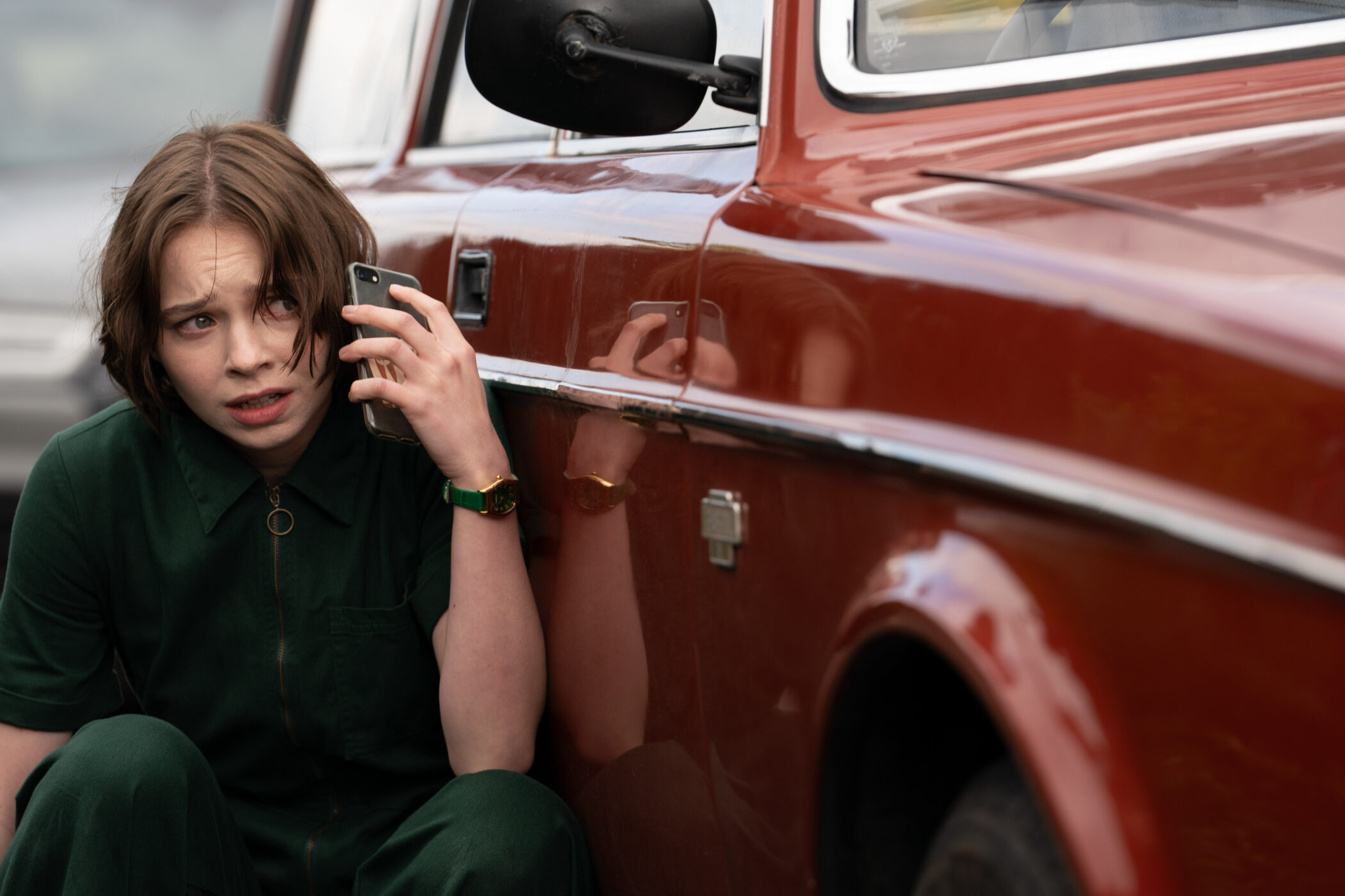 "Young person in green clothing crouches beside a red car, holding a phone to her ear and appearing worried or anxious, outdoors during daytime."