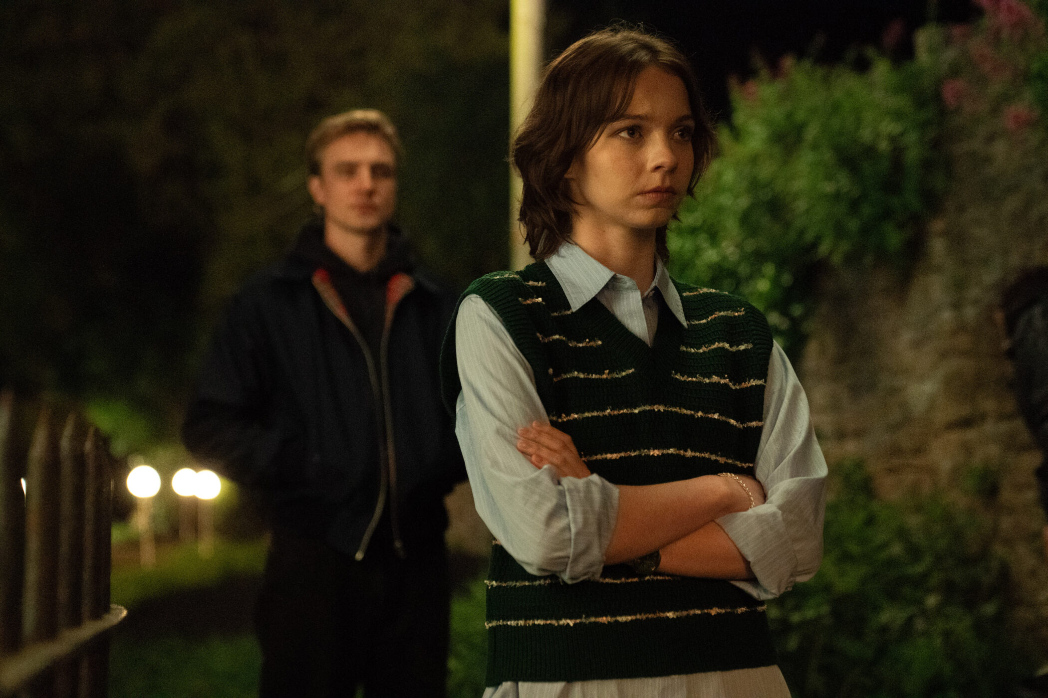 "Two young people standing outside at night, one in the foreground with arms crossed looking thoughtful, the other slightly blurred in the background. The environment is dimly lit with greenery and a stone wall visible."