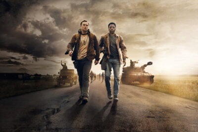 Cooper Hoffman and David Jonsson walk an open road under a storm sky, flanked by military vehicles and the rest of the column, in Francis Lawrence's The Long Walk.