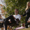 "A family of four, two adults and two kids, sit outdoors on folding chairs on a hillside, enjoying a sunny day with trees and autumn foliage in the background."
