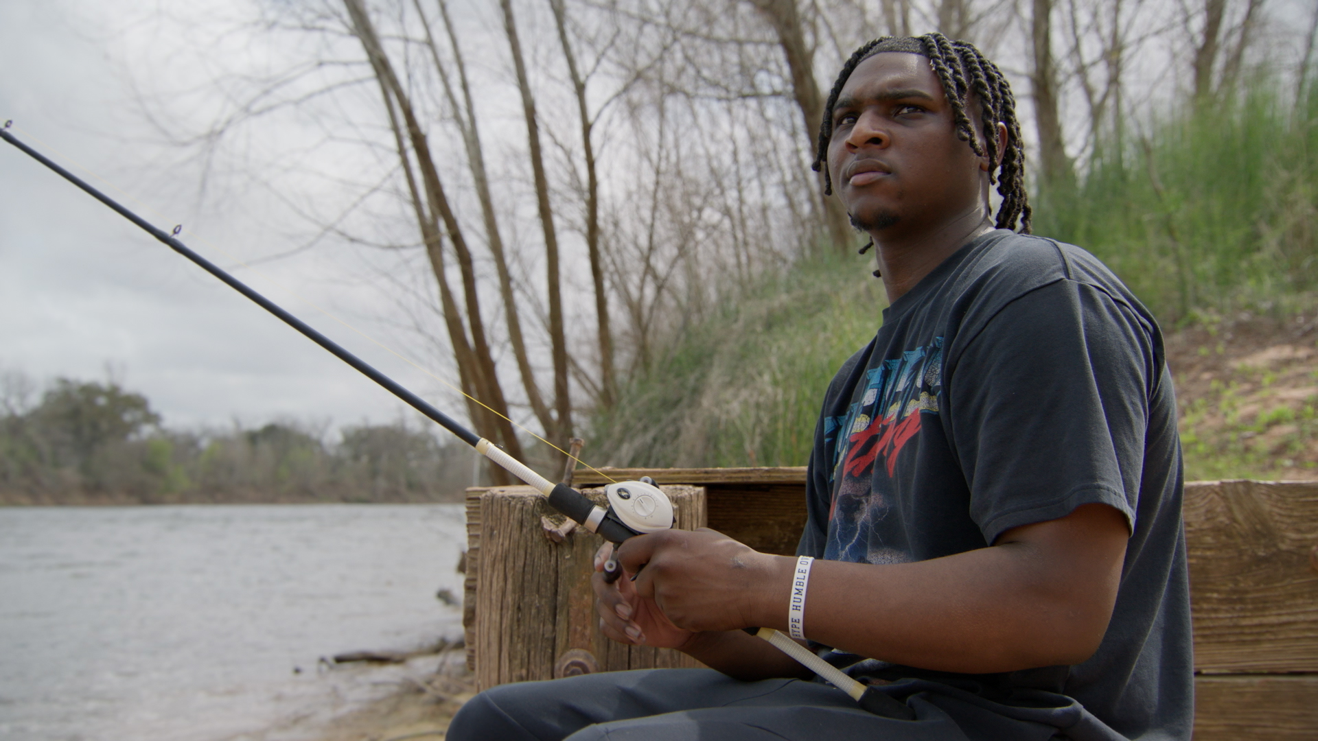 "Young man fishing by a riverbank, sitting on a wooden bench surrounded by trees and greenery on a cloudy day, holding a fishing rod and looking thoughtful."