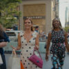 "Three women walk together in a city, smiling and carrying shopping bags, with tall buildings, a Bulgari store, and people in the background on a sunny day."
