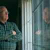 "Older man in plaid shirt stands indoors with arms crossed, gazing thoughtfully out a window. His reflection is visible in the glass, and the room is dimly lit, creating a contemplative, quiet atmosphere."