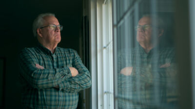 "Older man in plaid shirt stands indoors with arms crossed, gazing thoughtfully out a window. His reflection is visible in the glass, and the room is dimly lit, creating a contemplative, quiet atmosphere."
