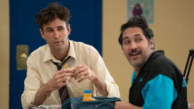 John Reynolds eats a packed school lunch beside a classmate in a high school cafeteria scene from the comedy Never Change!