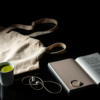 Editorial still life: a canvas tote bag with a matcha cup, an unmarked paperback and a single wired earbud laid out like catalogued evidence under a hard spotlight on dark glass.