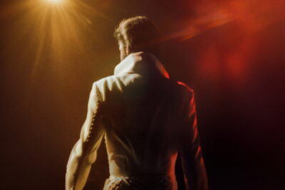 Elvis Presley in a high-collared jumpsuit, seen from behind under a stage spotlight, in a still from Baz Luhrmann's concert documentary EPiC: Elvis Presley in Concert.