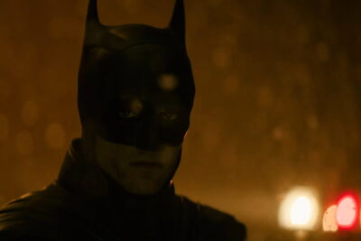Robert Pattinson as Batman in the cowl, rain-slicked Gotham night with police lights behind, from The Batman