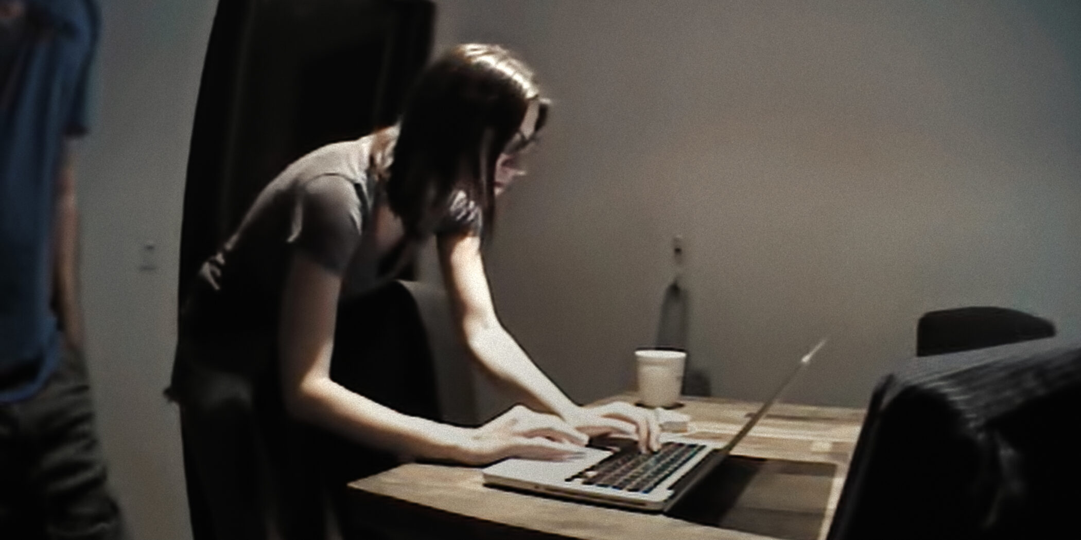 "Woman standing and typing on a laptop at a wooden table in a dimly lit room with a cup and chairs nearby."