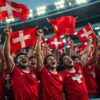 Football supporters dressed in red wave Swiss flags and celebrate together in a packed stadium stand under floodlights.