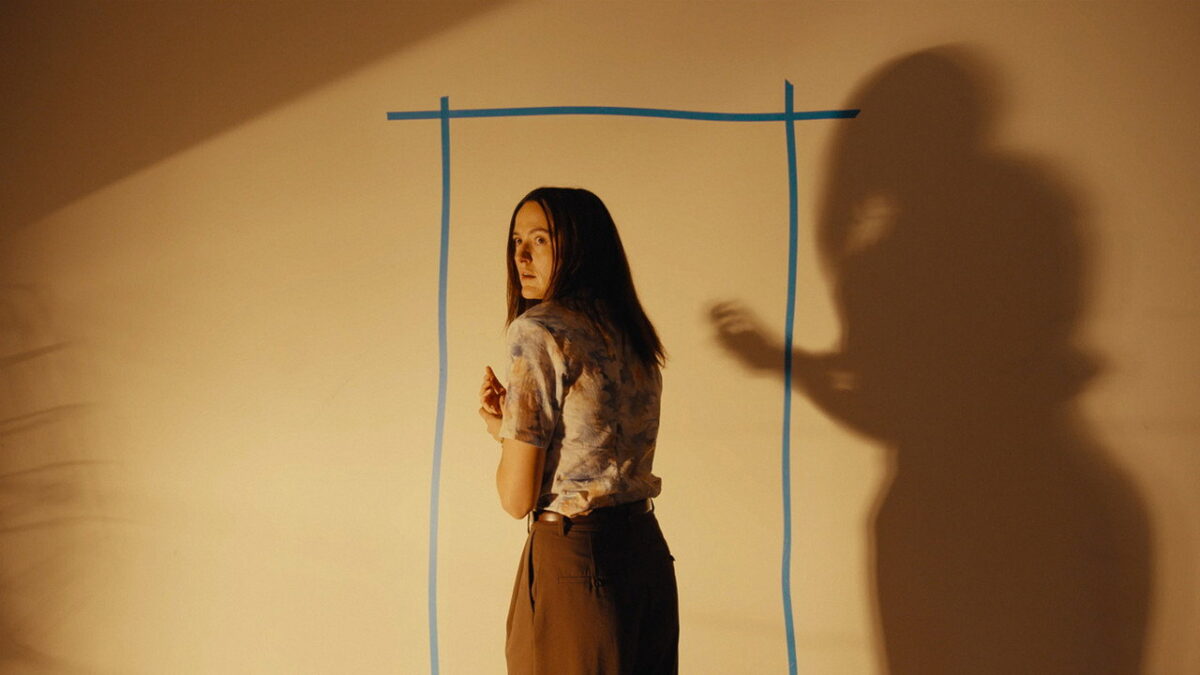 A woman stands before a blue-tape doorway outline in BACKROOMS, an ominous shadow looming beside her