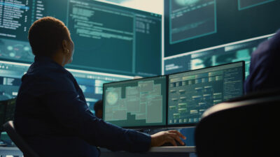 A cybersecurity analyst working in front of monitors showing code and network data