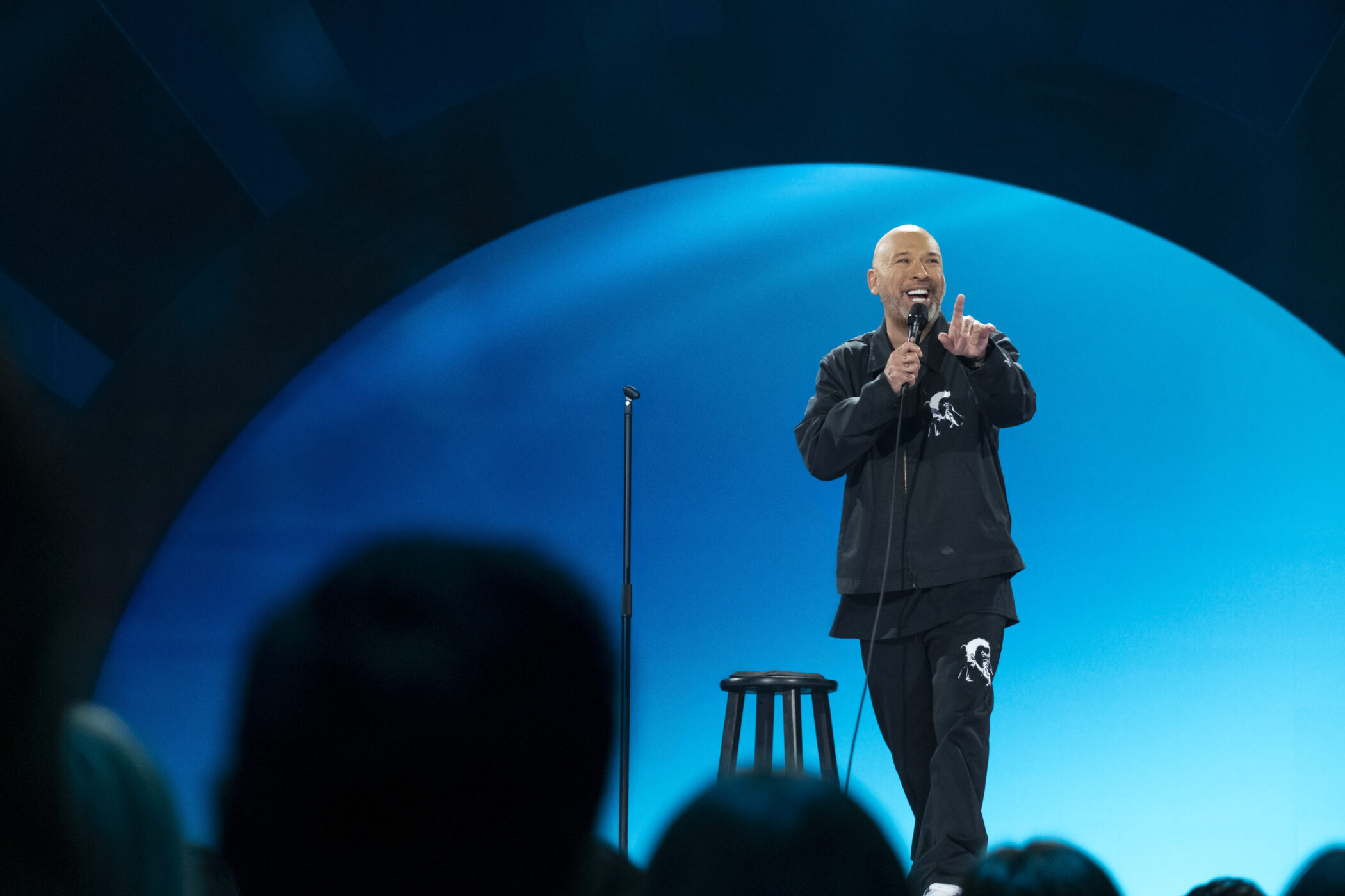 Jo Koy points at the audience in front of a blue circular backdrop in Blue in the Face