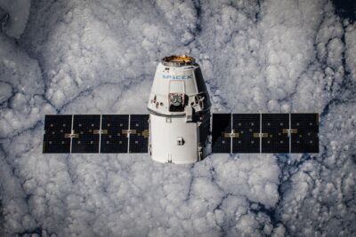 SpaceX Dragon spacecraft in orbit above Earth with solar panels extended and the SpaceX logo visible on the capsule hull