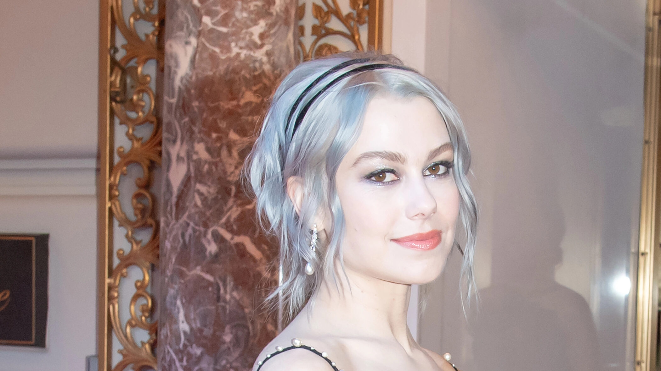 Phoebe Bridgers with pale blue hair held by a dark headband, wearing a sheer black dress with pearl beading, in an ornate gilded interior