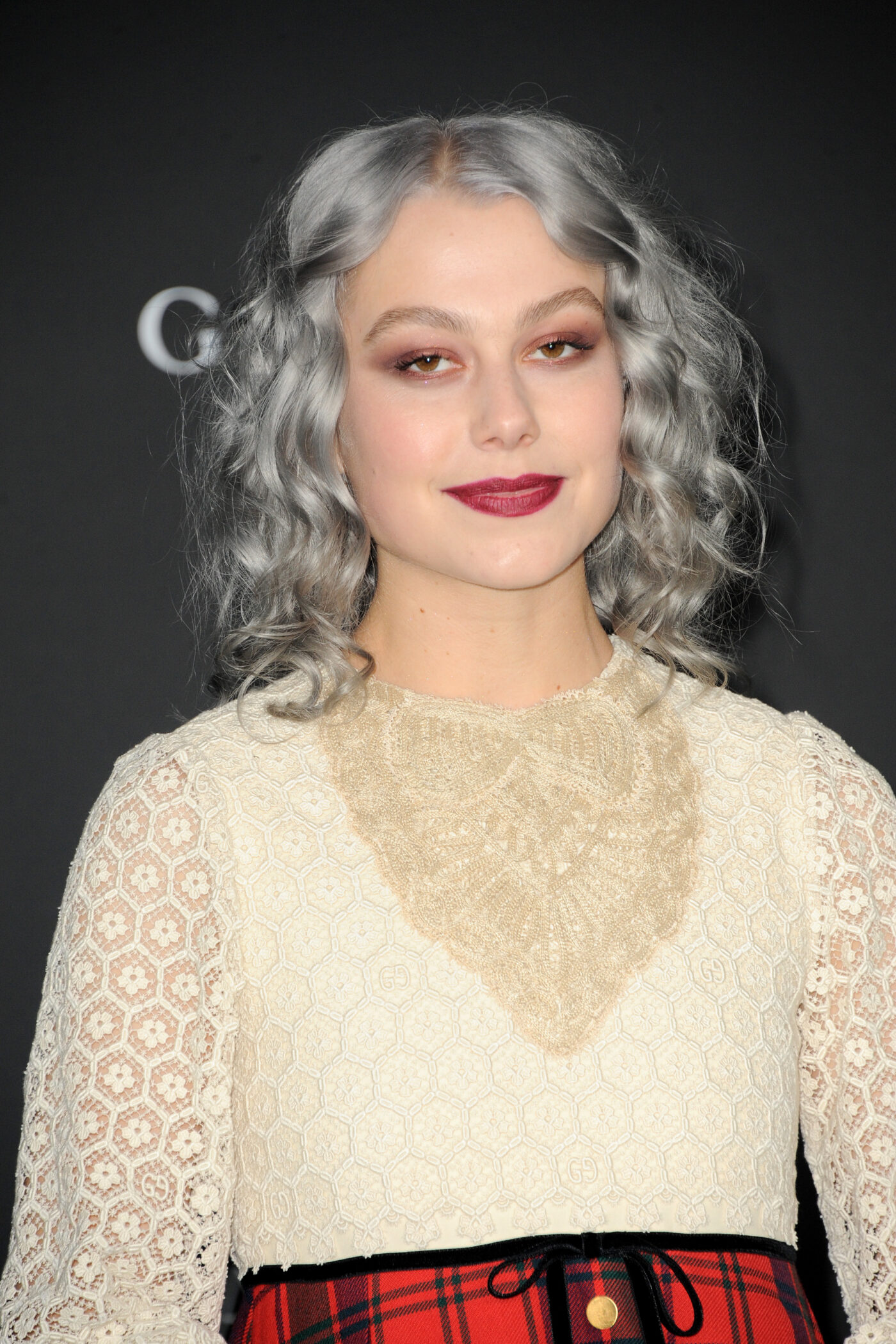 Portrait of Phoebe Bridgers with silver-grey curls, wearing a cream lace blouse over a red tartan skirt, against a dark backdrop
