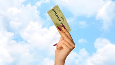 A hand holds up a golden ticket against a blue sky in Wonkas The Golden Ticket