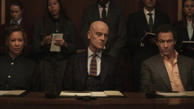 Dominic West, Nina Sosanya and Pip Torrens seated at a long conference table with lawyers standing behind them in a still from the legal drama War
