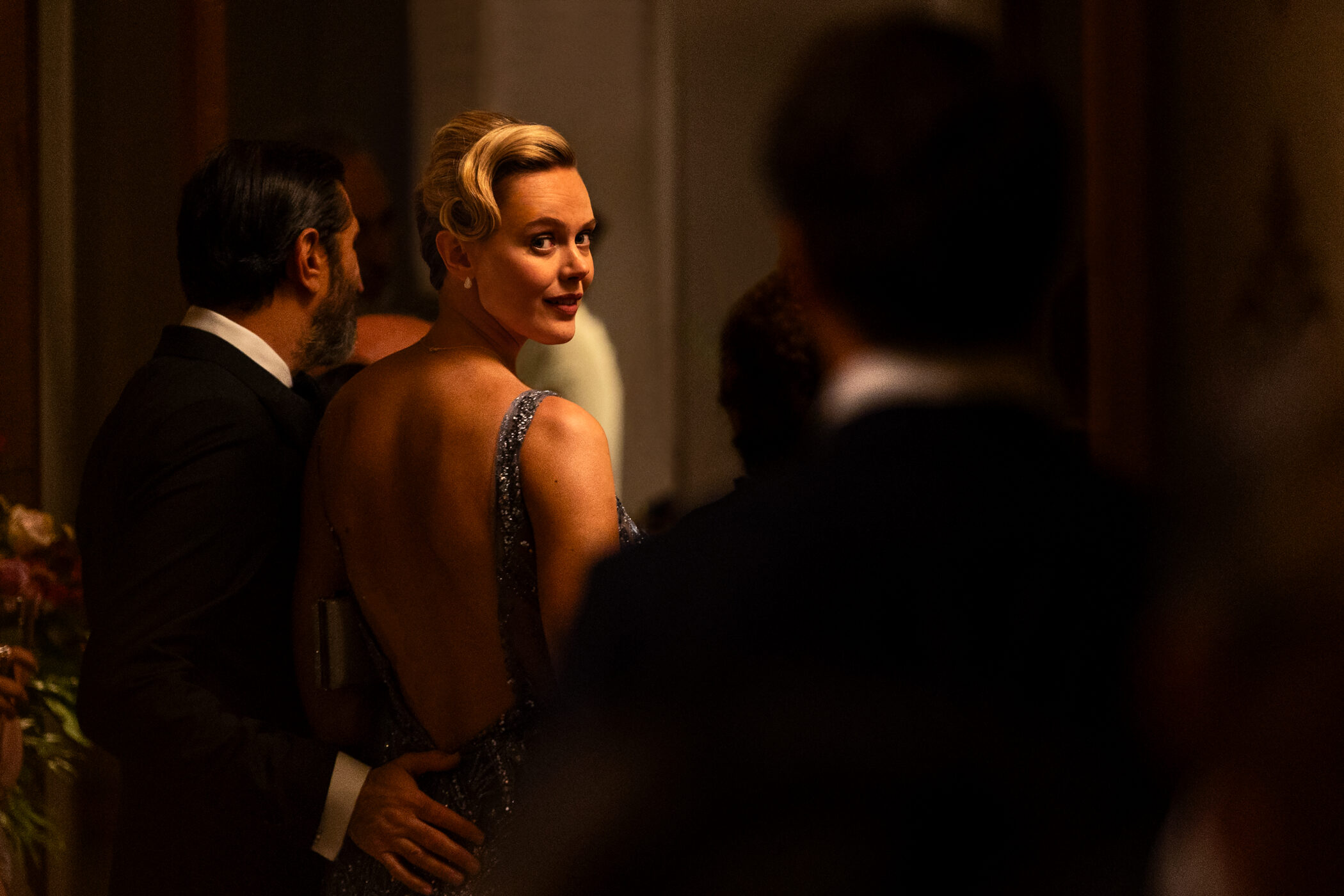 A woman in an open-back evening gown looks over her shoulder at a formal party in A Couple of Lies (Netflix)