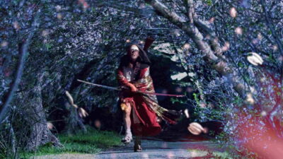 A candle-crowned, armored figure advances through night-lit blossom in Takashi Shimizu's The Village of Eight Gravestones.