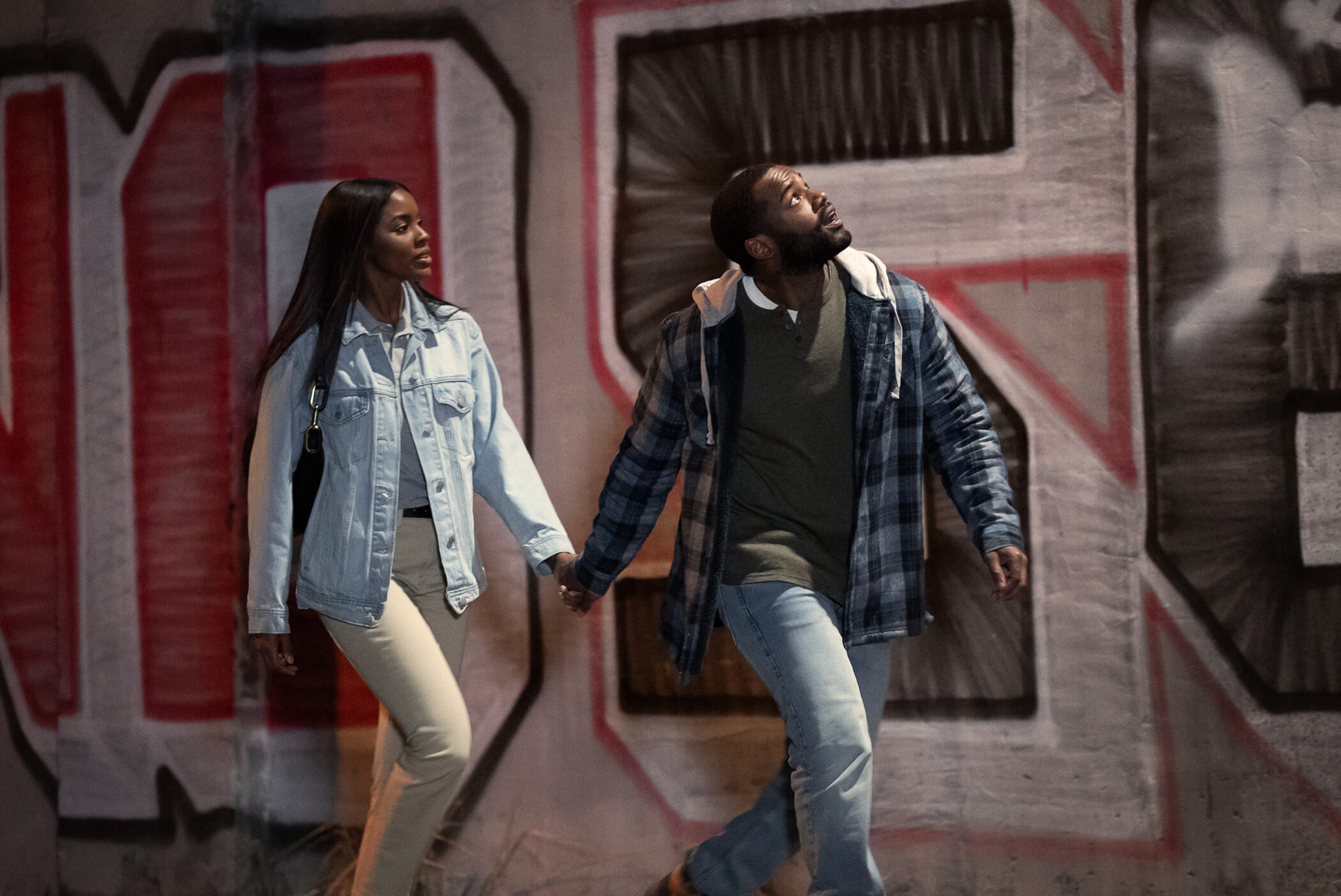 A couple walking hand in hand past a graffitied wall in Doing Life