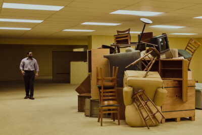 Chiwetel Ejiofor as Clark standing in an endless fluorescent-lit yellow room beside a heap of stacked furniture in Backrooms