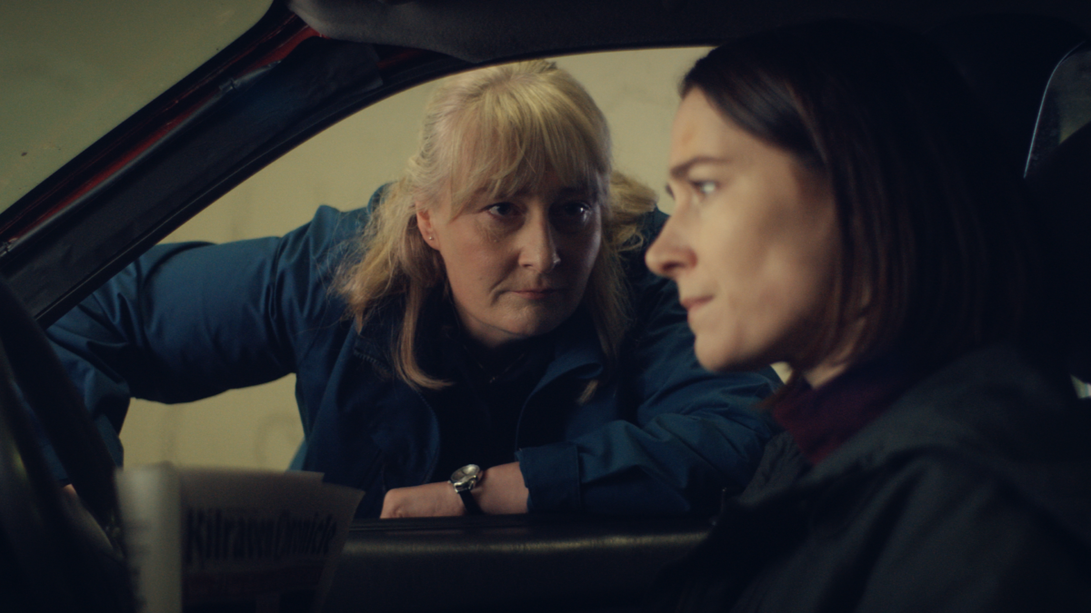 Scene from Obituary: a woman leans into a car window to speak to another woman in the Irish dark comedy series