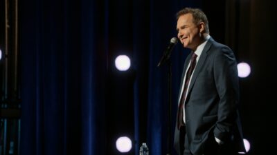 Norm Macdonald smiling at the microphone on a stand-up stage