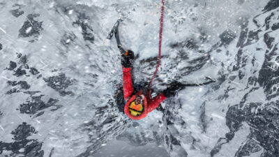 A climber in a red down suit and yellow goggles hangs from ice axes on a frozen wall in driving snow, in the key art for The Last First: Winter K2 on Apple TV