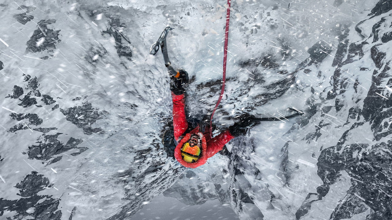 A climber in a red down suit and yellow goggles hangs from ice axes on a frozen wall in driving snow, in the key art for The Last First: Winter K2 on Apple TV