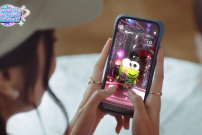 Hands holding a smartphone showing Keroppi dancing in Sanrio Kawaii Me Live rhythm game