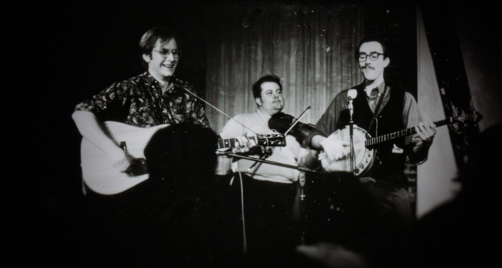 Black-and-white archive photo of a young Martin Mull playing guitar on stage with a fiddler and a banjo player