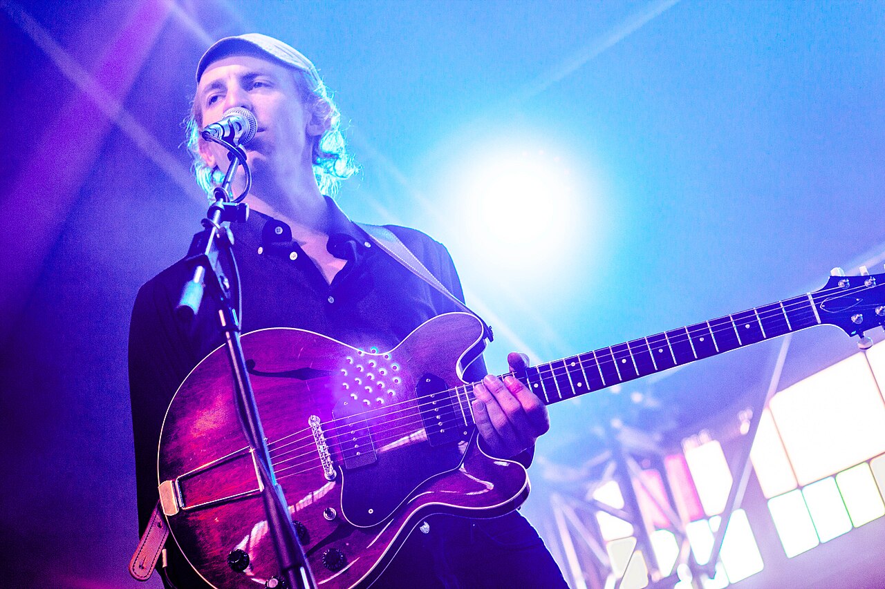 Buck Meek in a pale cap and dark shirt singing into a microphone while playing a hollow-body electric guitar on stage with Big Thief, under blue stage light
