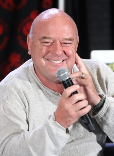 Dean Norris smiling and holding a microphone on a panel at Rose City Comic Con 2024 in Portland, Oregon, wearing a light grey sweater