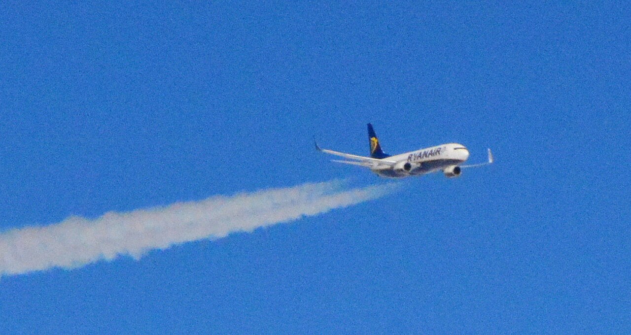 A Ryanair Boeing 737 climbing through a clear blue sky, trailing a long white contrail