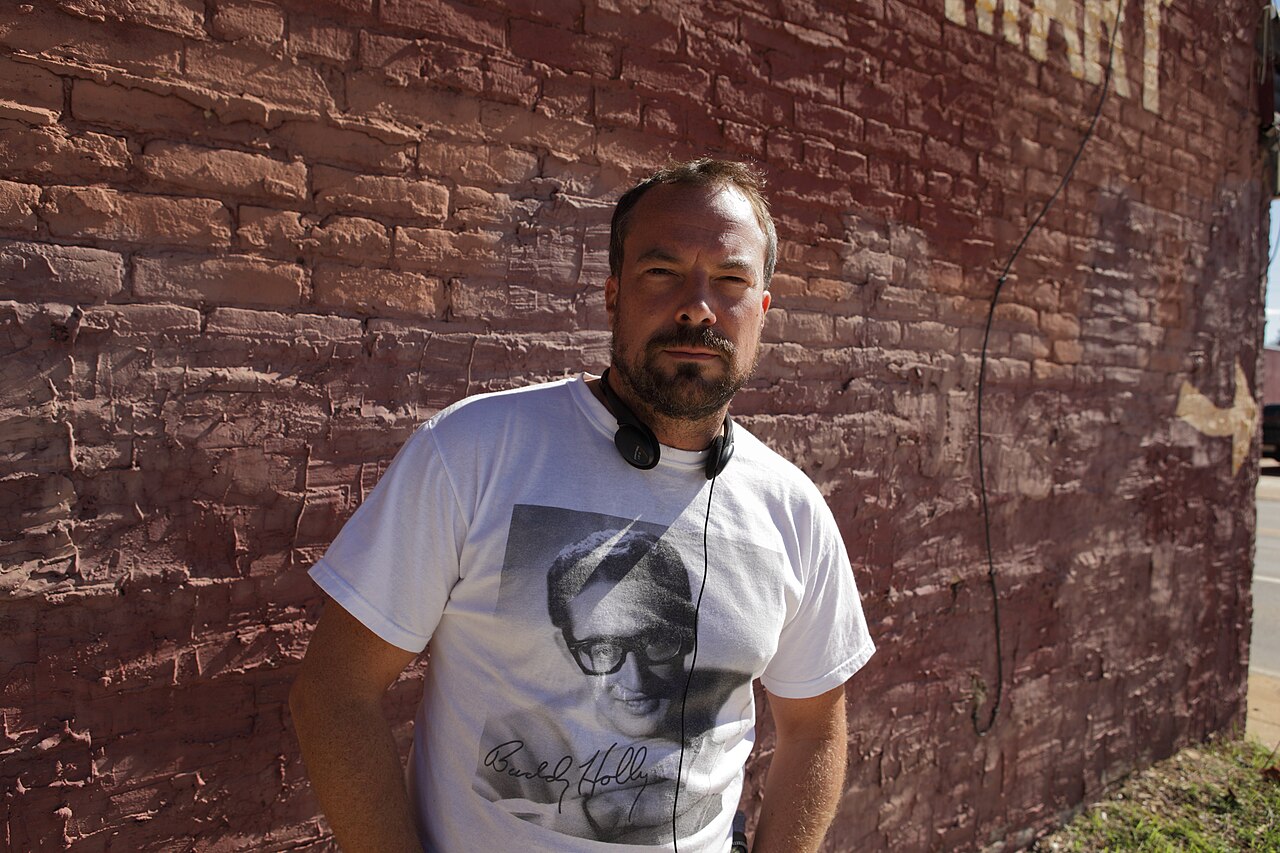 Simon Rumley in a white Buddy Holly T-shirt with headphones round his neck, standing against a red brick wall on a film set