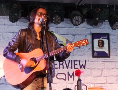 Bhuvan Bam in sunglasses and a leather jacket singing into a microphone while playing an acoustic guitar on stage