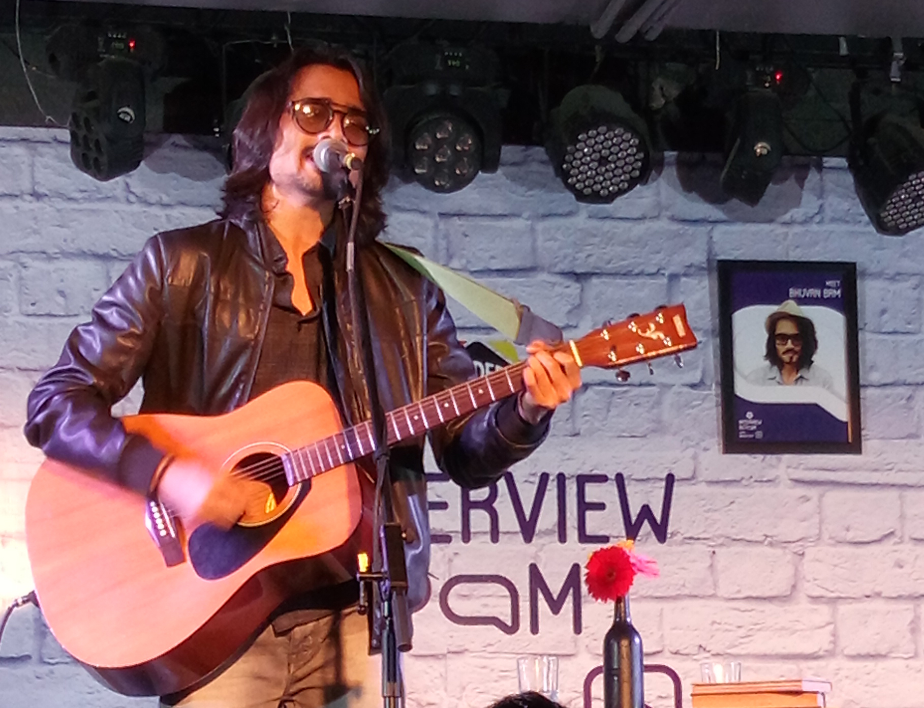Bhuvan Bam in sunglasses and a leather jacket singing into a microphone while playing an acoustic guitar on stage