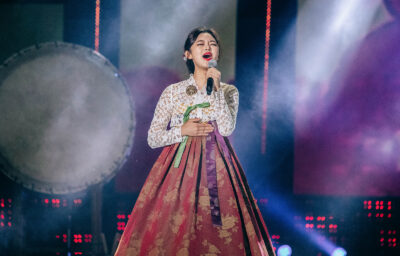 Jeong So-ri in a white lace jeogori and rose-patterned hanbok skirt, eyes closed, singing into a handheld microphone on a smoke-lit stage beside a large barrel drum