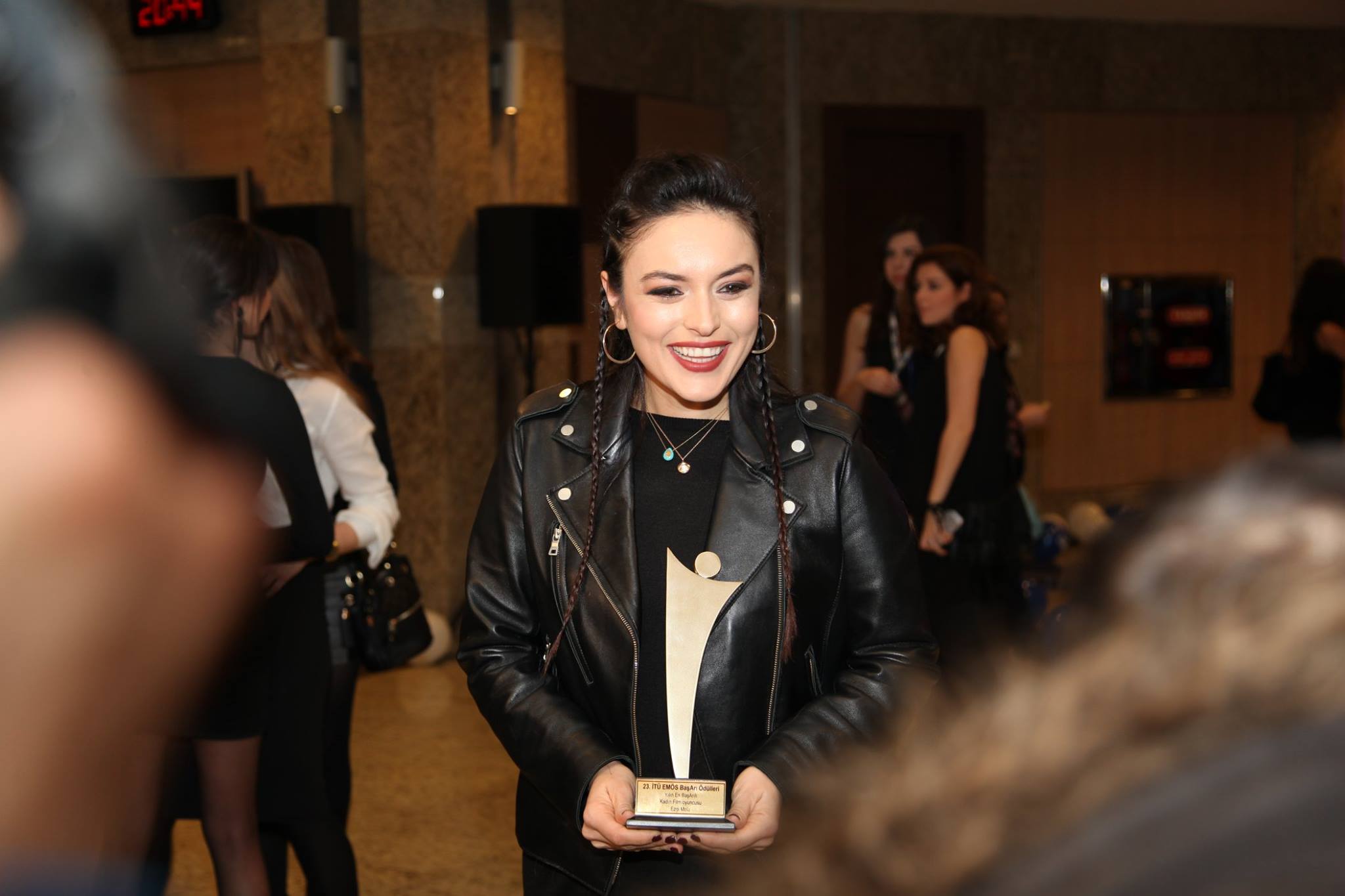 Ezgi Mola in a black leather jacket and braided hair, smiling and holding her İTÜ EMÖS best actress award, 2016