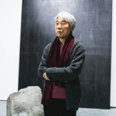Lee Ufan in a grey cardigan and burgundy scarf, arms folded, beside a boulder and a dark steel plate at his 2014 exhibition opening in Moscow