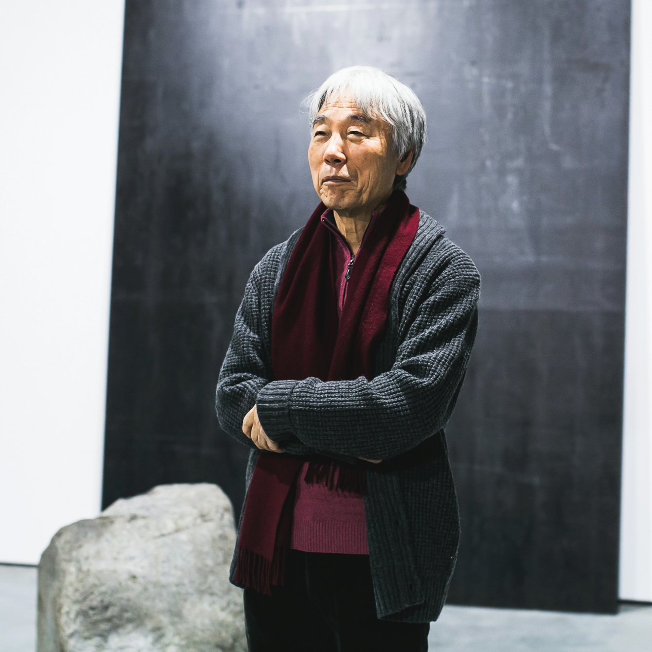 Lee Ufan in a grey cardigan and burgundy scarf, arms folded, beside a boulder and a dark steel plate at his 2014 exhibition opening in Moscow