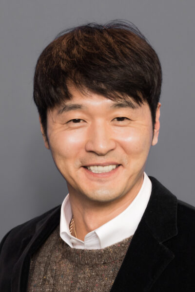 Lee Sung-jae, smiling, in a dark velvet jacket over a brown knit sweater and white shirt, at a Berlinale photo call in February 2018