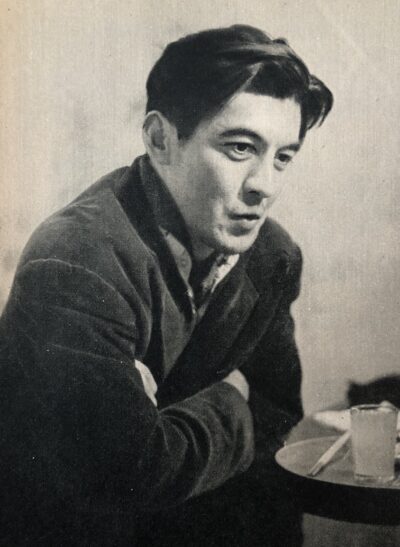 Black-and-white 1954 portrait of Rentaro Mikuni in a dark corduroy jacket, arms folded, seated beside a small café table