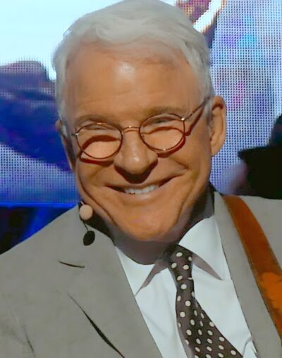 Steve Martin smiling on stage in round glasses and a grey suit during a 2017 concert in Concord, California