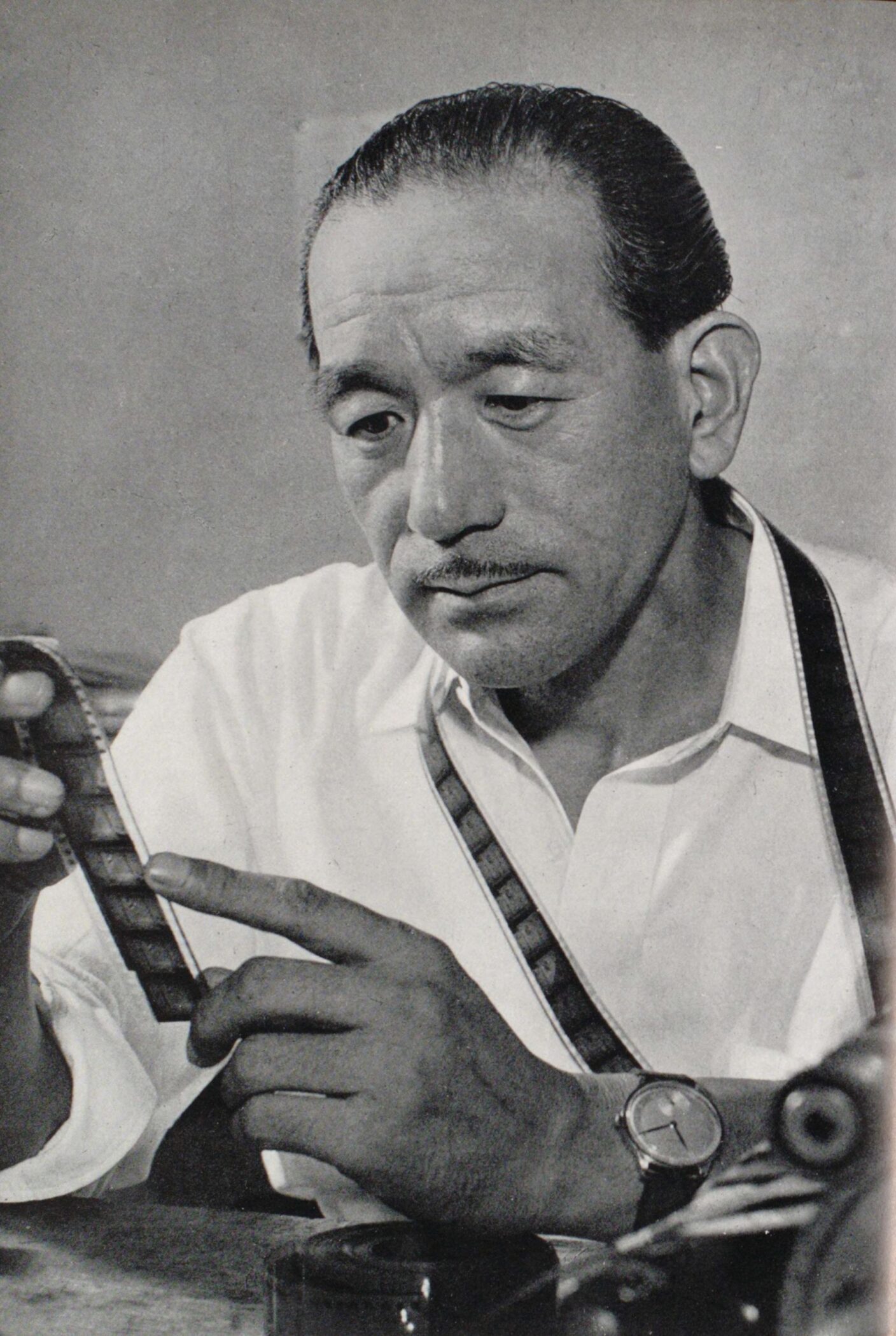 Yasujiro Ozu at a table examining a strip of film negative, with another strip hung around his neck, in 1953