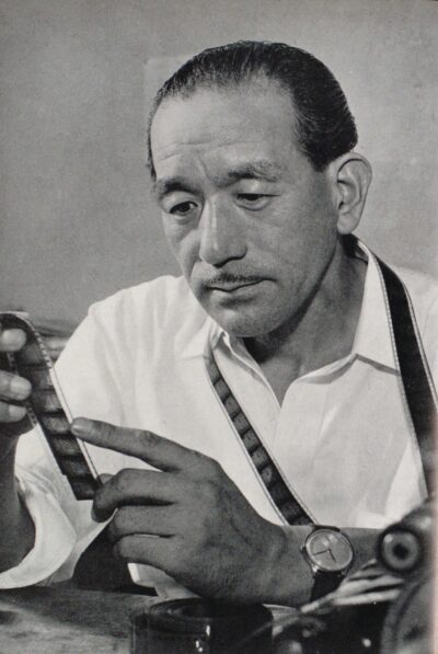 Yasujiro Ozu at a table examining a strip of film negative, with another strip hung around his neck, in 1953