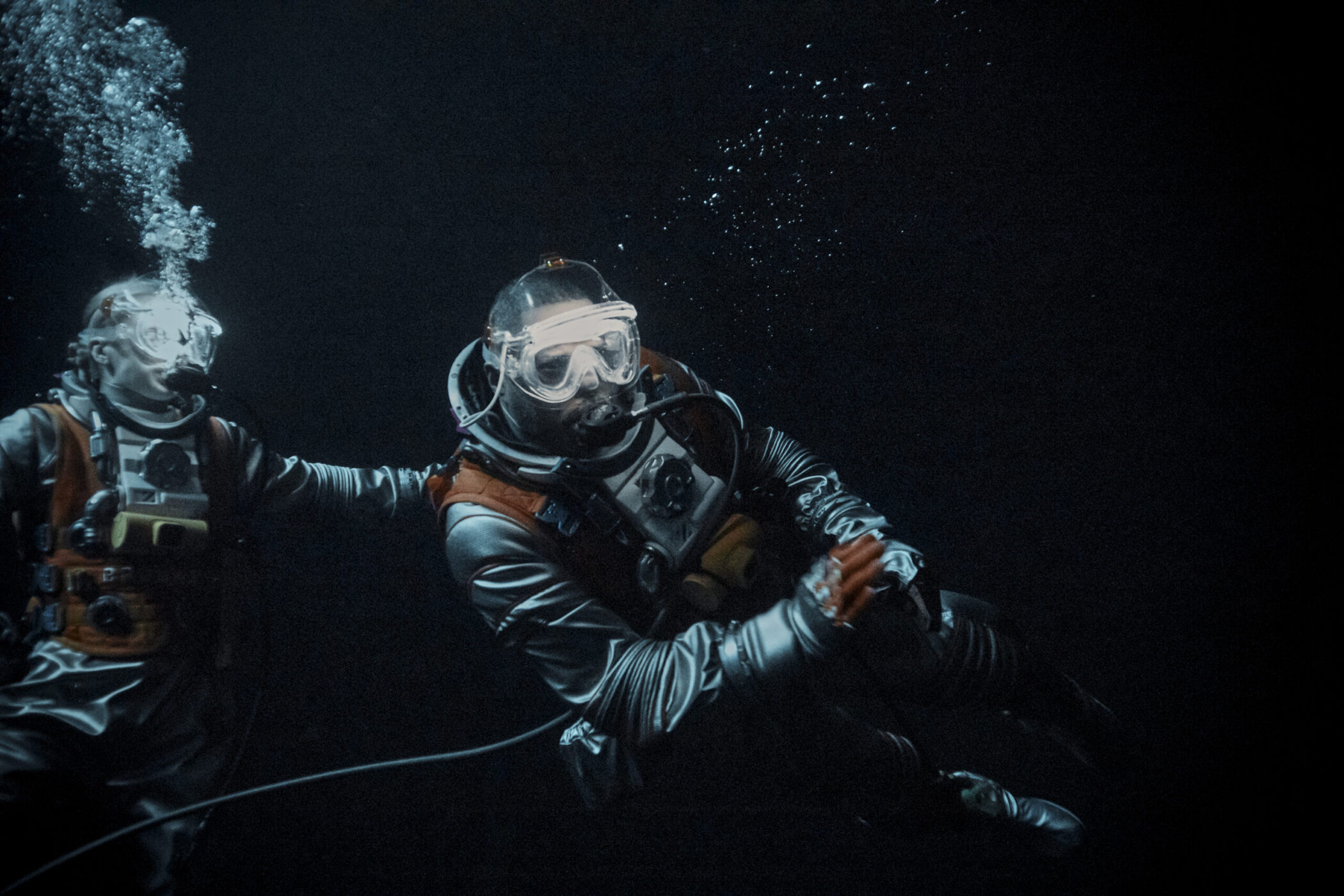 Two astronauts in diving masks swimming through dark water in Above the Below