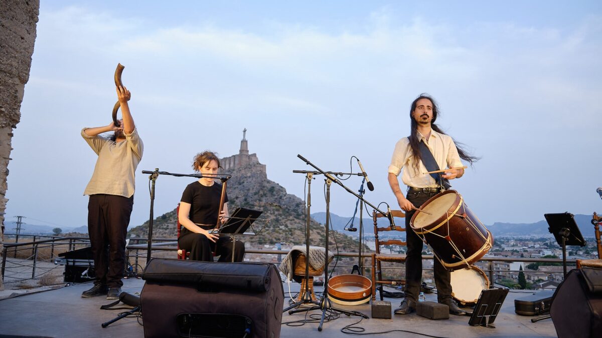 Al Tayr Ensemble performing outdoors at dusk with a horn, a recorder and a large drum beneath Monteagudo castle