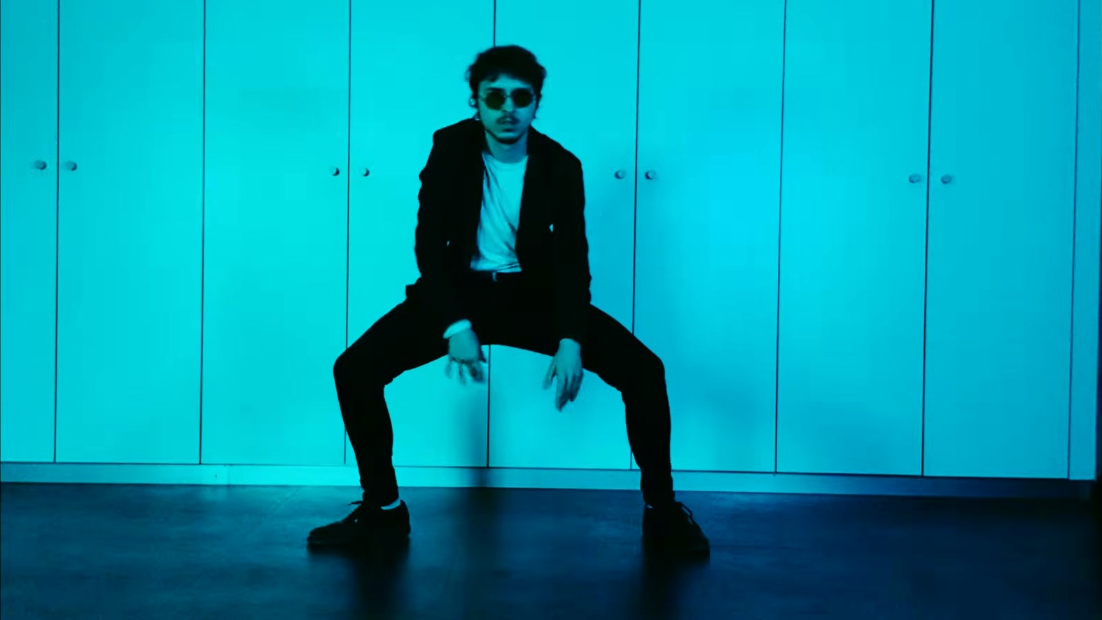 Albert Feiler in sunglasses and a black suit crouching in a wide dance stance against a cyan-lit wall